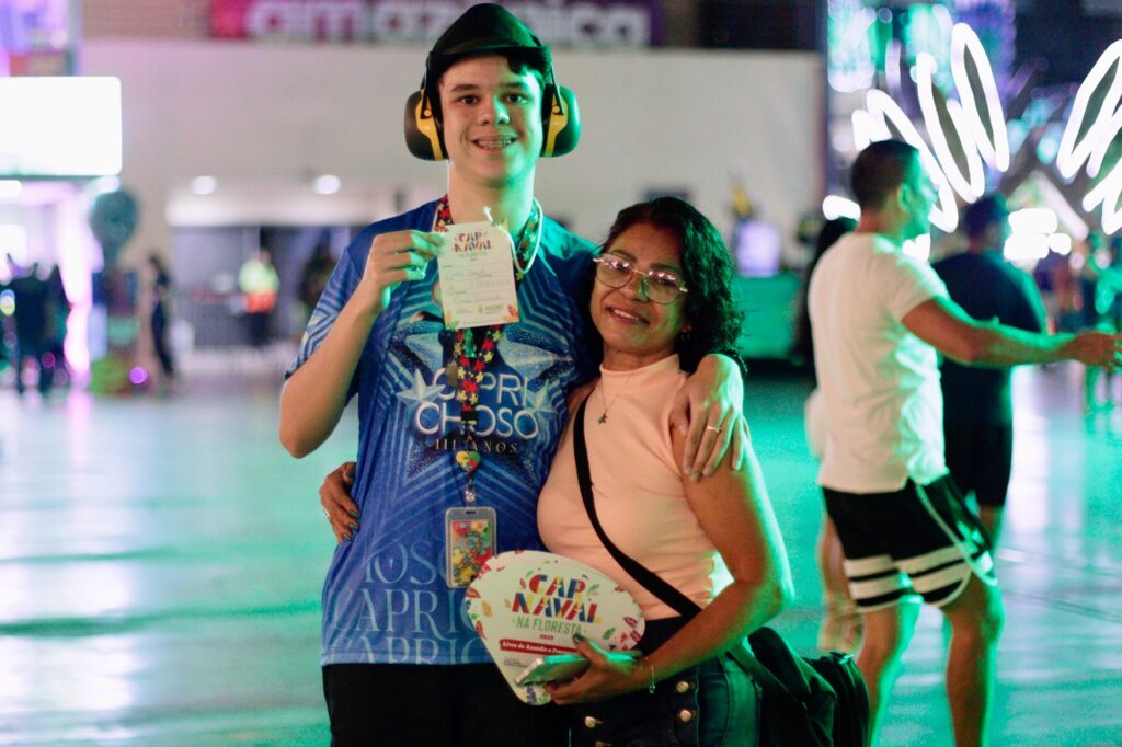 Imagem de uma mãe abraçada ao seu filho pcd, ambos sorrindo, ele está com um abafador, cordão autista e uma blusa do Caprichoso e a mãe com uma blusa básica