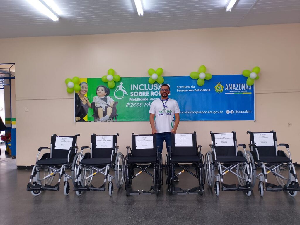 Servidor da secretaria junto com seis cadeiras de rodas enfileiradas para a entrega, com uma faixa do programa Inclusão sobre rodas e balões ao redor