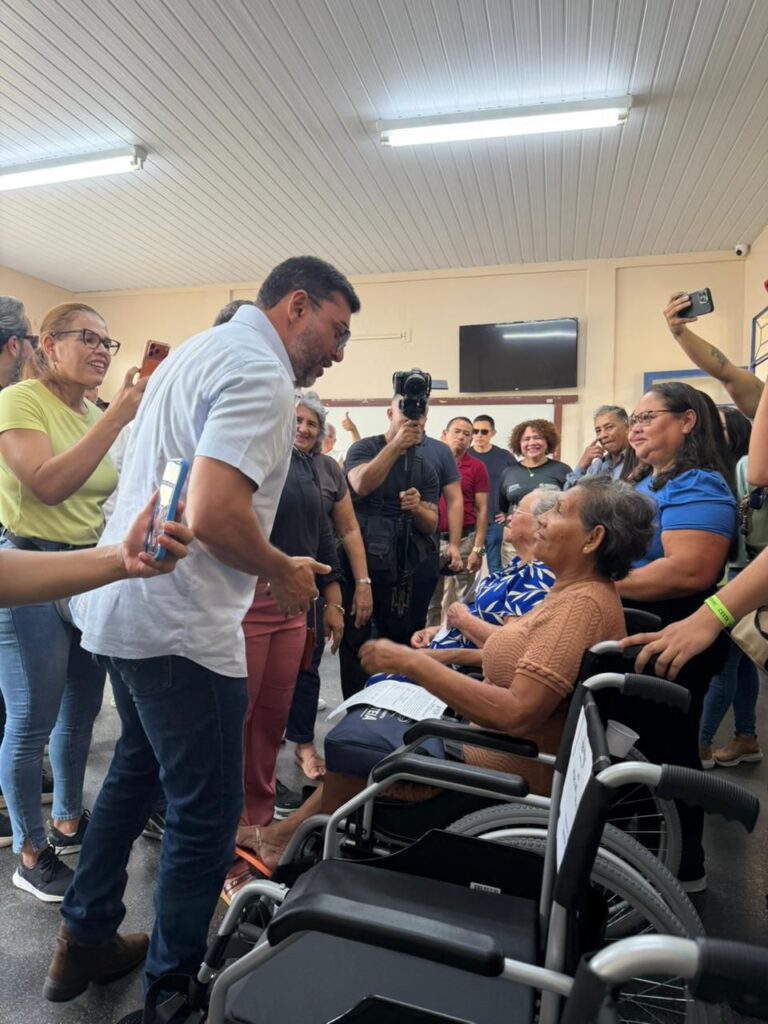 Governador Wilson Lima conversando com uma pessoa com deficiência em sua cadeira de rodas durante a entrega. Equipes de comunicação e familiares ao redor das cadeiras de rodas