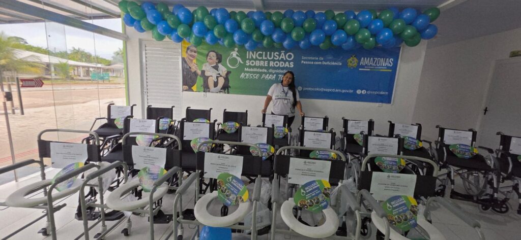 Servidora da sepcd com diversas cadeiras de rodas e de banho enfileiradas e organizadas para a entrega. Atrás dela há uma faixa do programa e balões pendurados