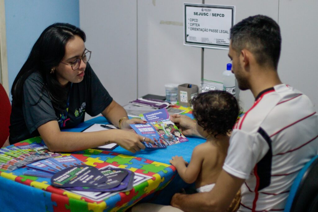 imagem da servidora em atendimento em uma ação, ela esta sentanda em uma mesa orientando um pai e um bebe a sua frente 