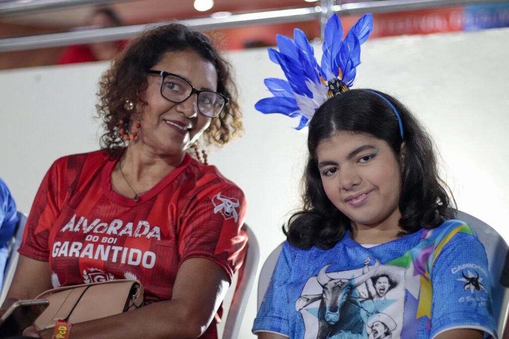 imagem de uma mãe e sua filha com deficiência no espaço acessível do bumbódromo, cada uma está torcendo para um boi: a mãe de vermelho, a filha de azul