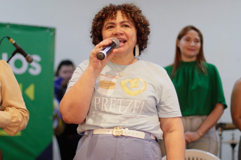 Imagem da secretária Selma Banes falando ao microfone, ela esta com uma blusa e calças azul claro e ao fundo está o painel do baile inclusivo