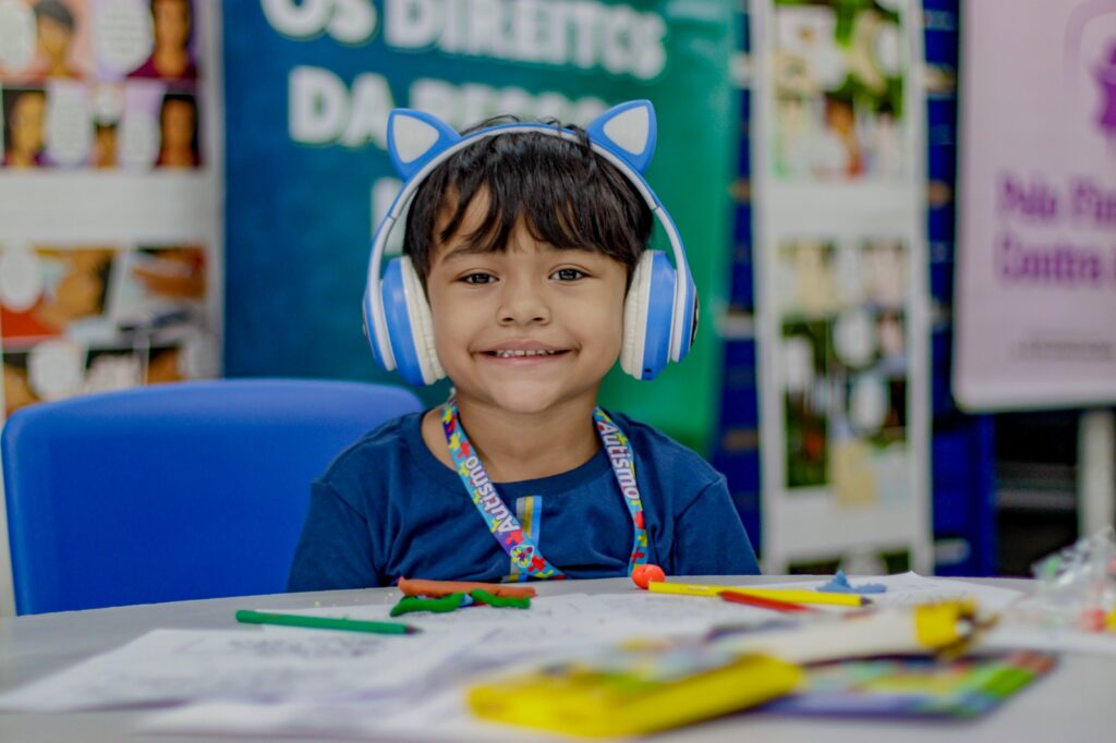 um menino sorrindo, vestindo um abafador azul e o cordão de quebra-cabeças no pescoço
