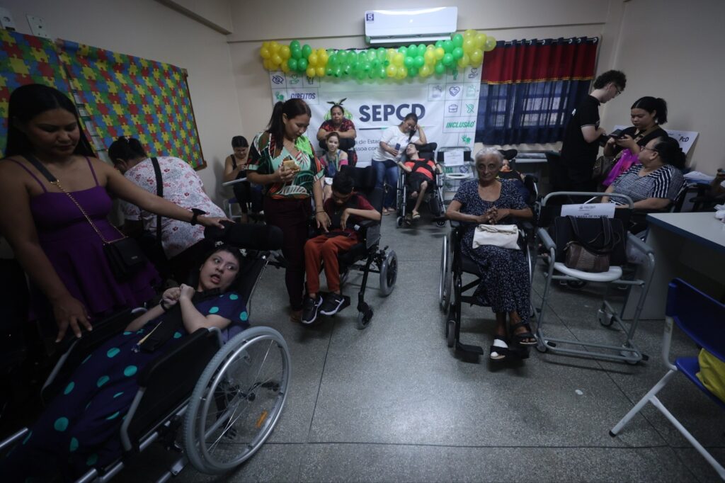 a imagem mostra várias pessoas em uma sala cheia de cadeiras de rodas e elas sentadas nelas, algumas são pcd, outros idosos e seus acompanhantes ao lado deles